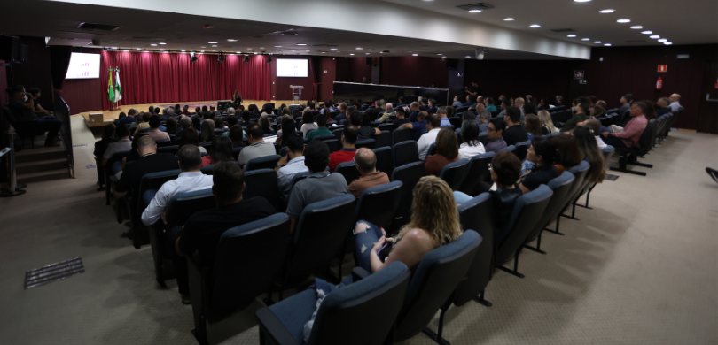 Fotografia da plateia de um auditório, sentada em cadeiras com estofado azul diante de um palco,...