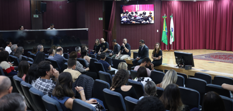 Fotografia de duas mulheres e quatro homens sentados na beira do palco de um auditório de frente...