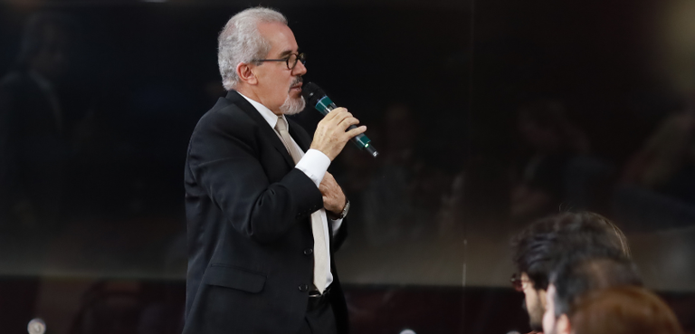 Na foto, um homem de terno preto, camisa social branca e gravata bege caminha em direção a plate...