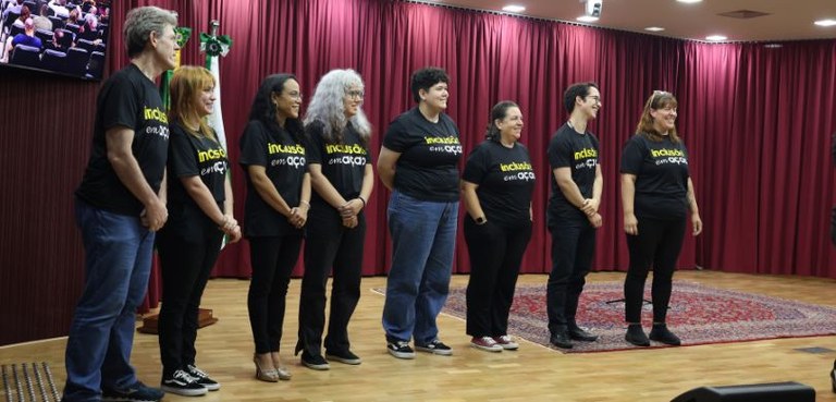 Homens e mulheres posam sorrindo lado a lado no palco do auditório do TRE-PR. As pessoas vestem ...