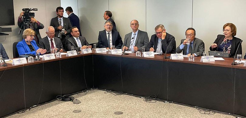 Fotografia de uma mesa em formato de ‘L’ com diversas autoridades, entre homens e mulheres. Sobr...