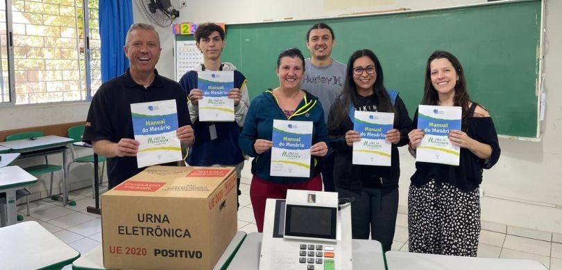 Em uma sala com paredes brancas e janelas de vidro, observam-se três homens e três mulheres posa...