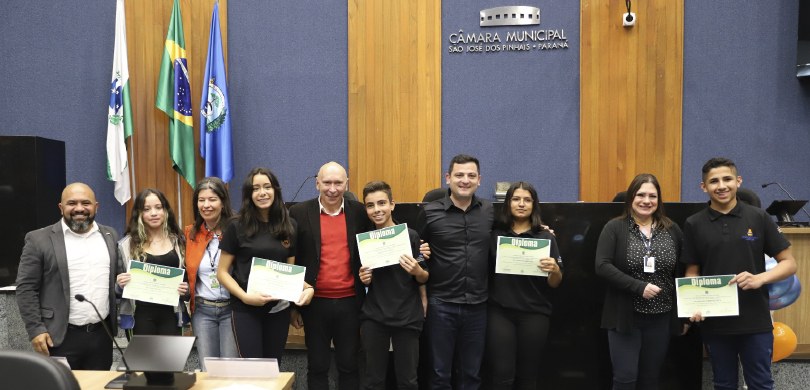 Fotografia de 10 pessoas, entre homens e mulheres, posando sorrindo no plenário da Câmara Munici...