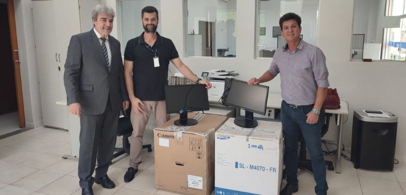 Fotografia de três homens posando sorrindo ao redor de duas caixas de papelão sobre as quais há ...