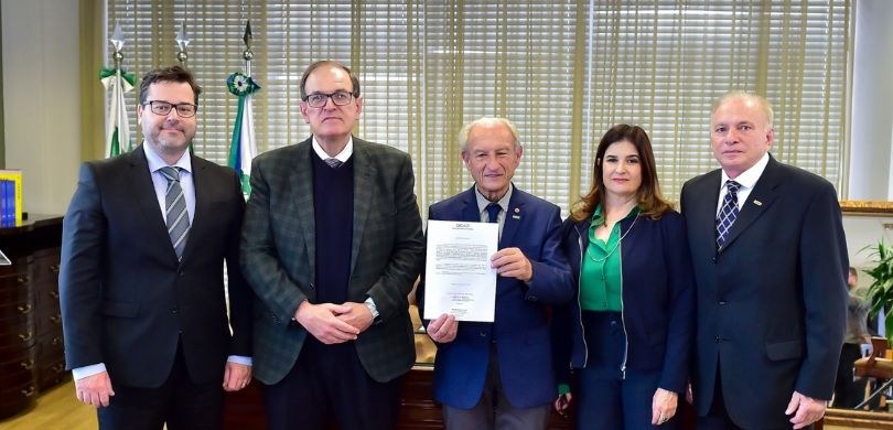 Fotografia de quatro homens de terno e uma mulher, que veste camisa verde e calça e jaqueta azui...