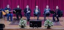 Fotografia de um grupo de seis homens sentados em cadeiras em um palco de auditório tocando inst...