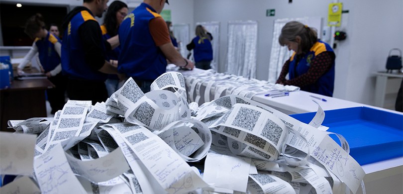 Fotografia das servidoras e servidores da Justiça Eleitoral mexendo nos boletins de urna. Ao fun...