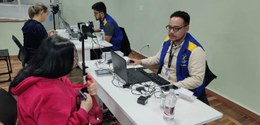 Fotografia de dois homens vestindo coletes da Justiça Eleitoral, nas cores azul e amarela. Eles ...