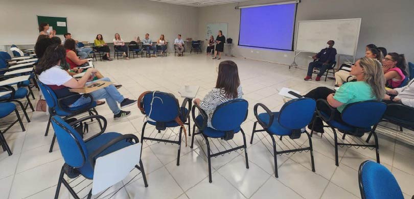 Em uma sala de aula com cadeiras azuis, várias pessoas estão sentadas em círculo. Ao fundo, um t...