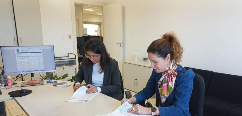 Foto em que aparecem duas mulheres sentadas em frente a uma mesa clara, assinando documentos. No...