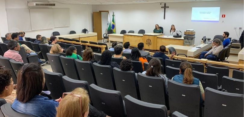 Foto em que aparecem em primeiro plano pessoas sentadas de costas em poltronas escuras dentro de...