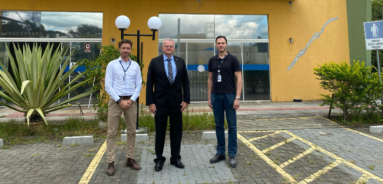 Fotografia de três homens posando sorrindo em frente ao Fórum Eleitoral, que tem portas de vidro...