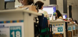 Fotografia de várias mulheres sentadas diante de mesas brancas, sobre as quais há computadores e...