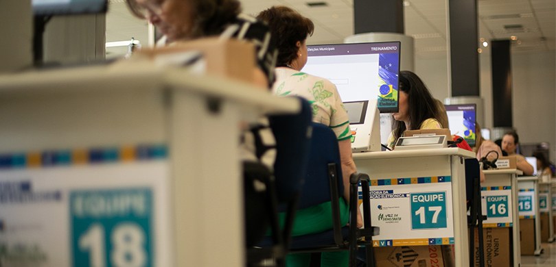 Fotografia de várias mulheres sentadas diante de mesas brancas, sobre as quais há computadores e...