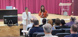 Fotografia de duas mulheres em pé falando diante de uma plateia, que aparece sentada em cadeiras...