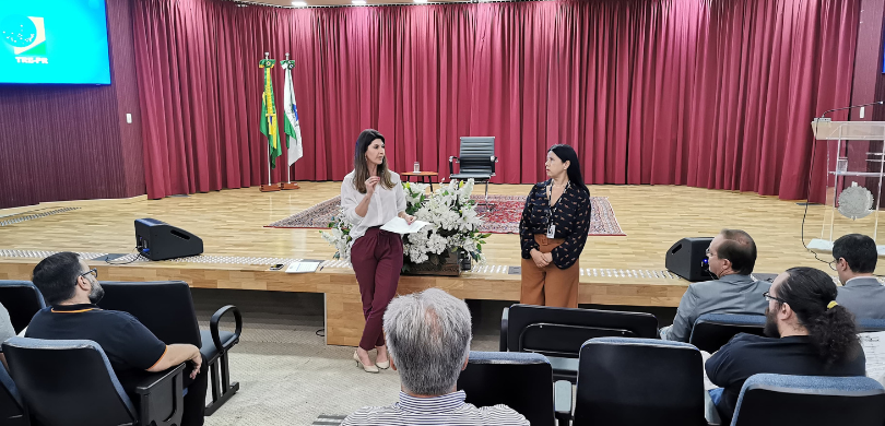 Fotografia de duas mulheres em pé, falando diante de uma plateia sentada em cadeiras com estofad...