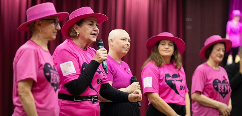 Cinco mulheres vestindo camisetas rosa (quatro delas com chapéus rosa) observam a plateia enquan...