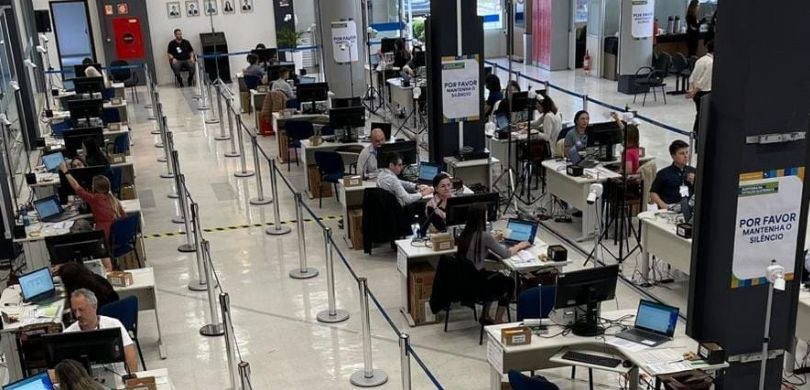 Foto tirada de cima em que aparecem várias pessoas sentadas em frente a tela de computadores sob...