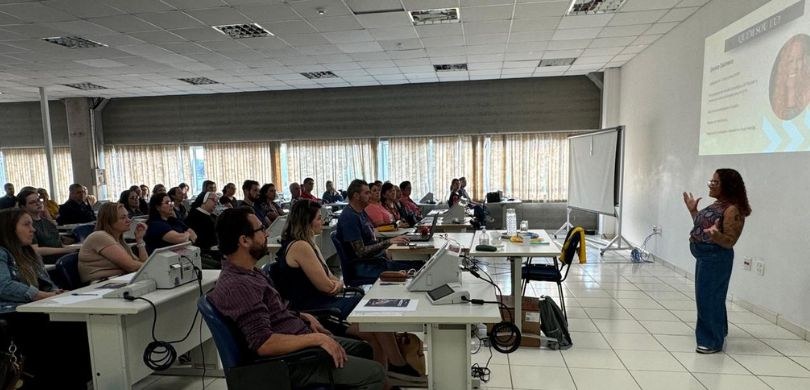 Foto em que aparecem, do lado esquerdo, de perfil, várias pessoas sentadas diante de mesas branc...