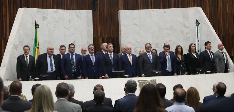Fotografia de homens e de mulheres atrás de uma bancada branca, no Plenário da ALEP. Em primeiro...