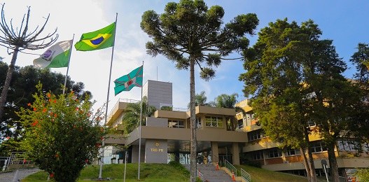 Fotografia da fachada de um prédio público, cercado de árvores. Há as bandeiras do Paraná, Brasi...