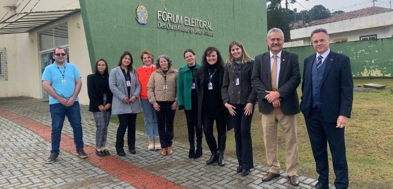 Fotografia de três homens e sete mulheres posando sorrindo em frente à fachada de um Fórum Eleit...