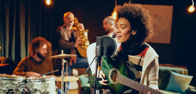 Fotografia de quatro pessoas tocando instrumentos. Em destaque, uma jovem mulher negra canta enq...