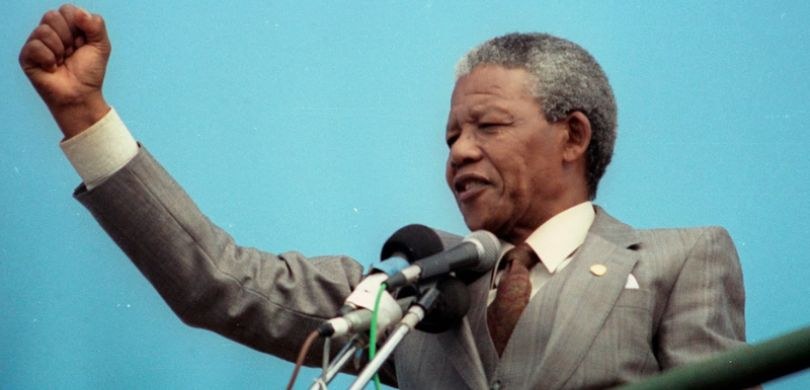 Fotografia de Nelson Mandela, um homem negro de cabelos curtos grisalhos, diante de microfones. ...