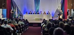 Fotografia de um palco de auditório sobre o qual se vê uma mesa de autoridades composta por três...