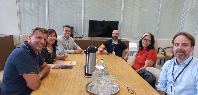 Imagem em que quatro homens e duas mulheres posam para foto, sentados ao redor de uma mesa de ma...