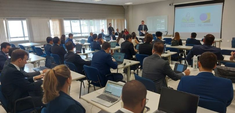 Foto em que aparecem em primeiro plano, sentado de costas em uma ampla sala de aula, juízas e ju...