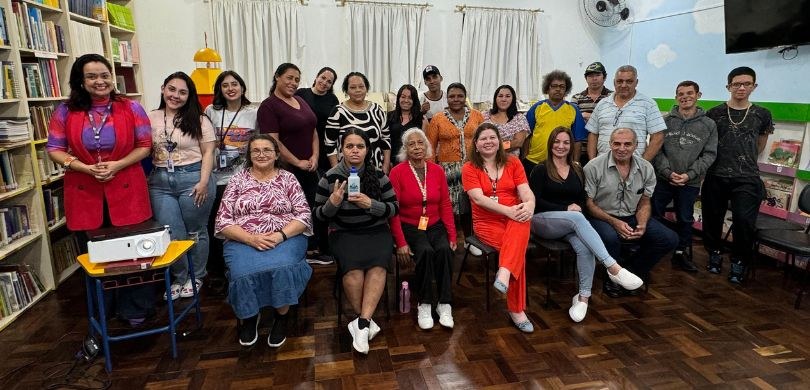 Em uma sala de aula, várias pessoas, entre homens e mulheres, sorriem para a foto. À frente de u...