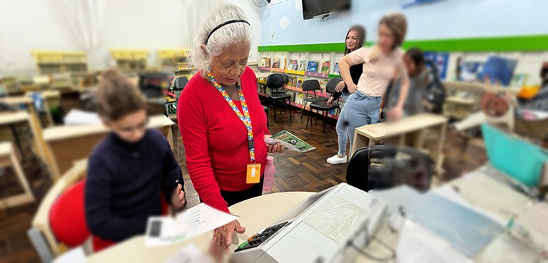 Foto de uma senhora ladeada por uma criança e três mulheres que a observam votar em uma urna ele...