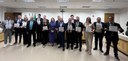 Fotografia de diversas pessoas posando sorrindo, a maioria segura em mãos um certificado branco....