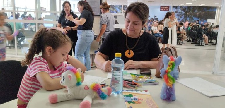 Em primeiro plano, do lado esquerdo, uma menina de cabelos claros desenha em uma folha de papel ...