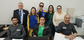 Fotografia de oito pessoas, quatro homens e quatro mulheres, posando sorrindo em uma sala com pa...