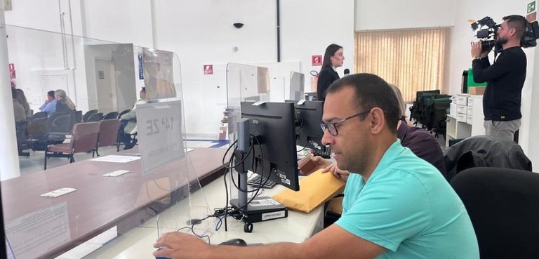 Foto em que aparece em primeiro plano, de lado, um homem usando óculos e uma camisa azul sentado...