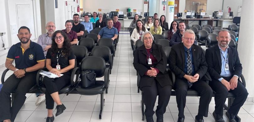 Foto em que aparecem várias pessoas sentadas em longarinas pretas, separadas por um pequeno corr...