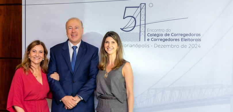 Fotografia de um homem de terno, ao centro, e duas mulheres posando sorrindo diante de um banner...