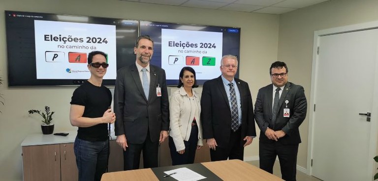 Fotografia de quatro homens e uma mulher posando sorrindo, em uma sala com paredes brancas. Ao f...