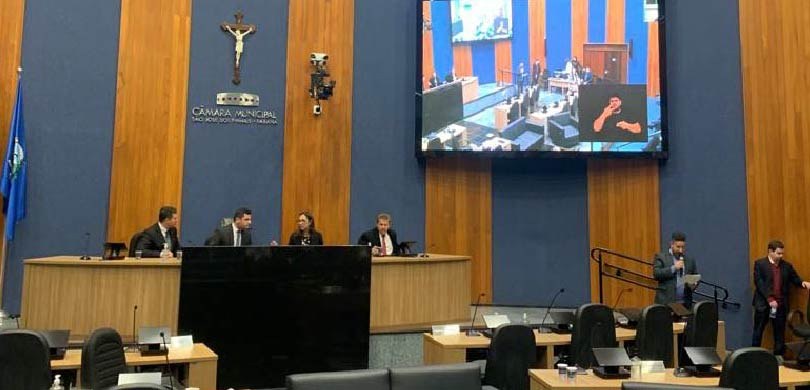Fotografia do plenário da Câmara Municipal. Portanto, há uma mesa de autoridades, uma televisão,...