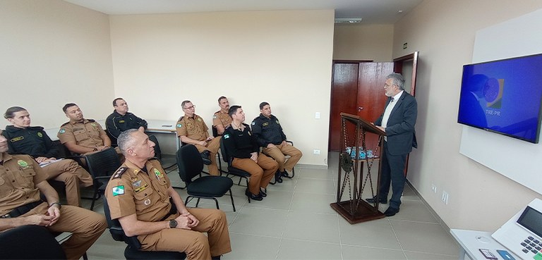 Fotografia de uma sala, com um homem falando a frente em um púlpito. Pessoas, que vestem a farda...