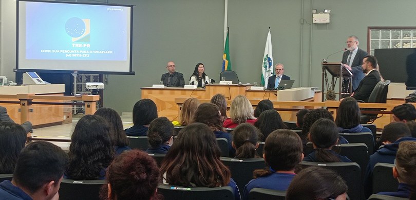 Fotografia de uma sala, com um homem vestindo terno discursando em um púlpito. Outras três pesso...