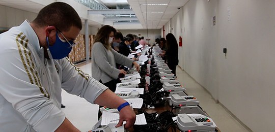 Fotografia de um corredor. Ao centro, há uma bancada com urnas eletrônicas sendo manipuladas por...