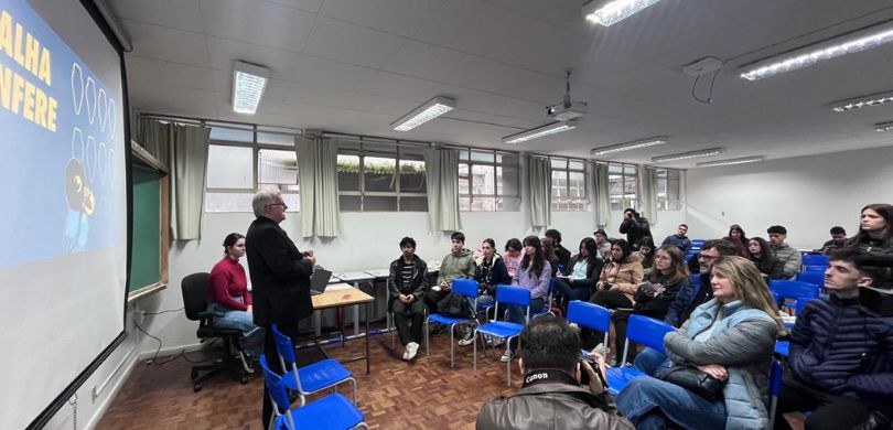 Fotografia de uma sala de aula. Do lado esquerdo, vê-se um homem, vestindo terno preto, apresent...