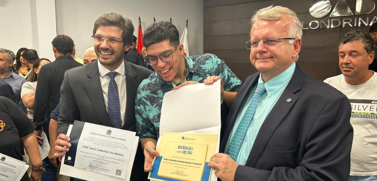 Fotografia de três homens, dois de terno e um com uma camisa estampada azul, posando sorrindo. U...