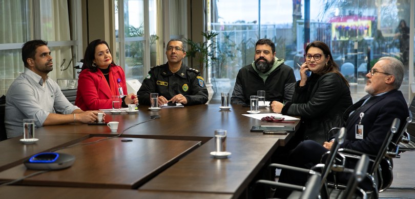 Foto em que aparecem sentados ao redor de uma mesa de madeira duas mulheres e quatro homens. Ao ...