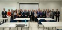 Fotografia de dezenas de pessoas, entre homens e mulheres, posando sorrindo em uma sala com pare...