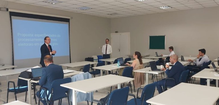 Fotografia de diversas pessoas, entre homens e mulheres, sentadas em carteiras azuis diante de c...