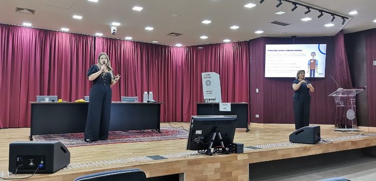 Fotografia de uma mulher, que veste roupas pretas, falando em um microfone em um palco de um aud...